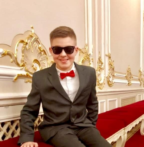 На фото Николай сидит на красной банкетке в красивом зале. Николай одет в темно-серый костюм и белую рубашку, а еще темные очки и красную бабочку.