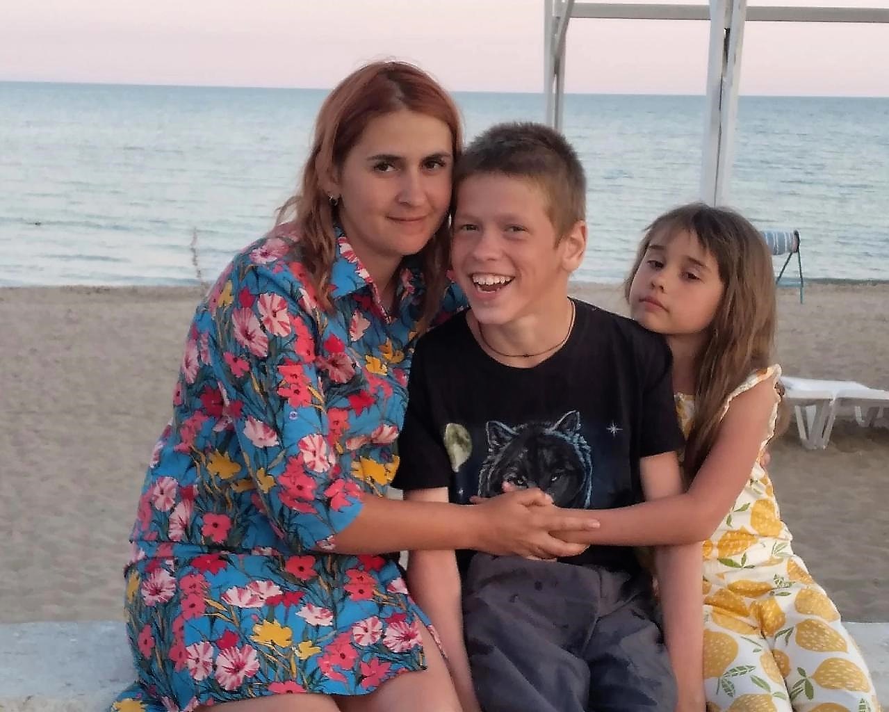 На фото Владислав сидит на берегу моря. Рядом с ним его мама и младшая сестра.