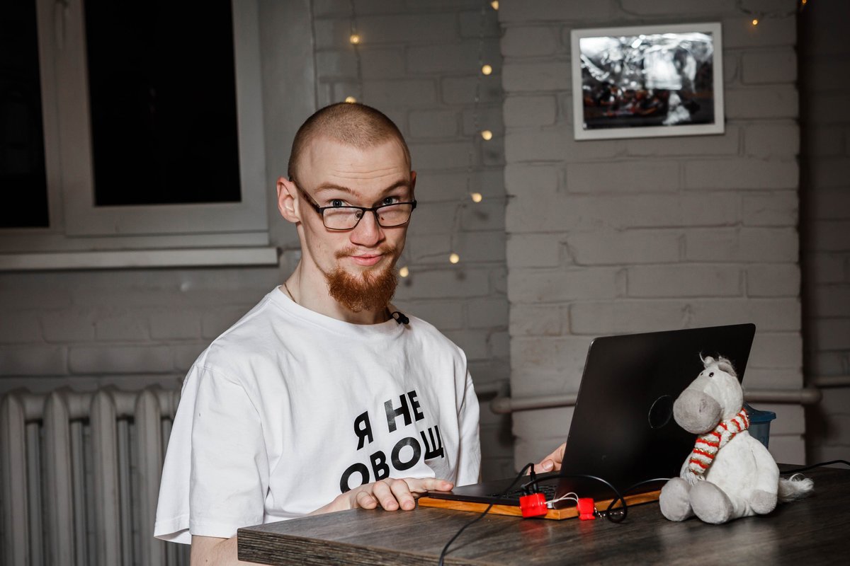 На фото Иван сидит за рабочим столом. На столе стоит ноутбук, за ноутбуком сидит мягкая игрушка серого цвета. У Ивана на белой футболке надпись: "Я не овощ".